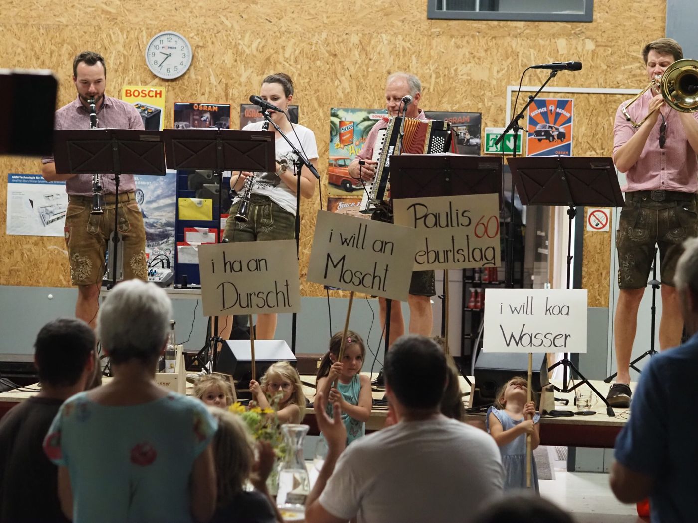 Die Hausmusik spielt auf einer Bühne, im Publikum halten Kinder Schilder mit „i ha an Durscht“ und „i will koa Wasser“.